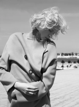Black and white portrait of a blonde woman wearing a tailored coat outdoors by the coast.