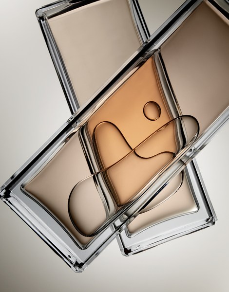 Close-up of two transparent cosmetic compacts with beige and tan foundation, stacked on a neutral background, reflecting light and shadow.