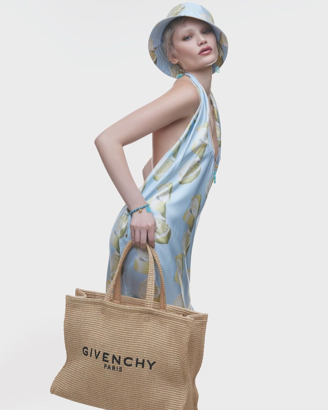Fashion model poses in a lemon-print dress and matching bucket hat, holding a straw tote bag against a plain studio background.