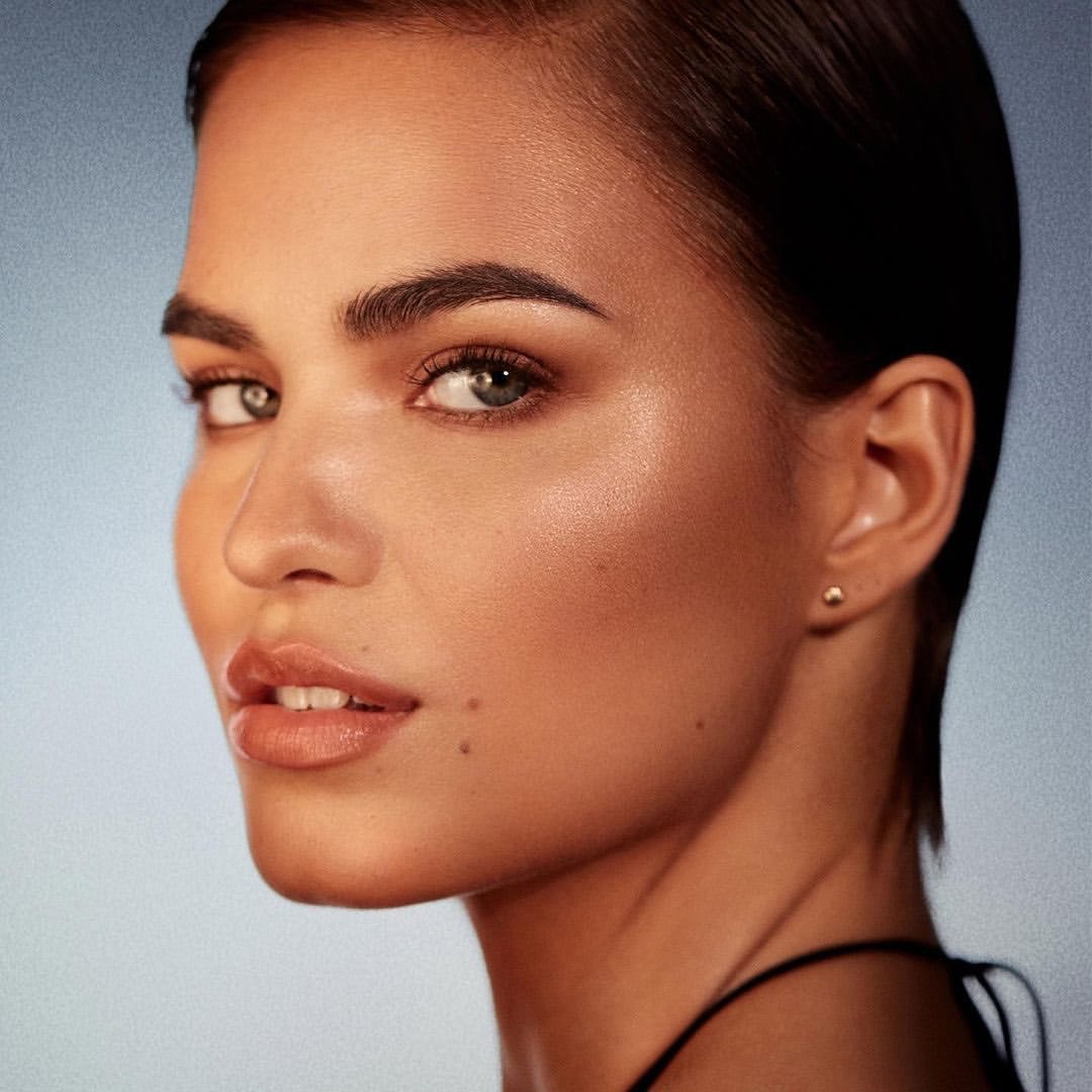 Close-up portrait of a woman with slicked-back dark hair, wearing glossy neutral lipstick against a subtle gradient blue background.