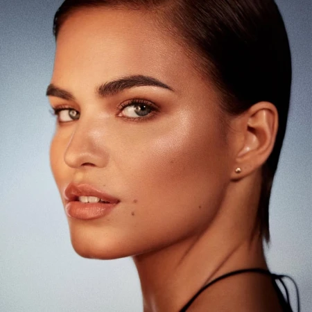 Close-up portrait of a woman with slicked-back dark hair, wearing glossy neutral lipstick against a subtle gradient blue background.