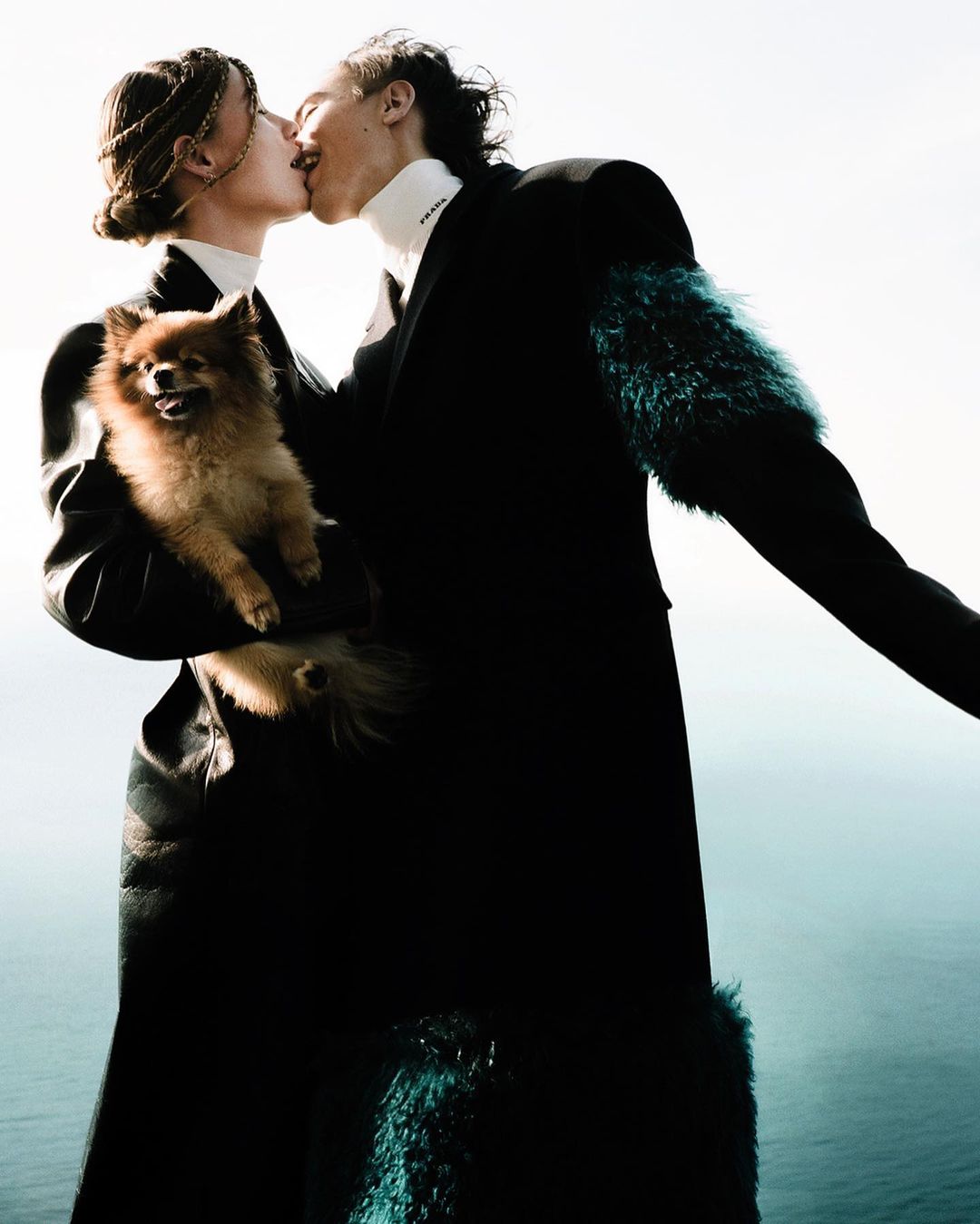 Two people kiss while holding a Pomeranian dog in an outdoor setting with a sea background, showcasing fashion and a touch of nature.