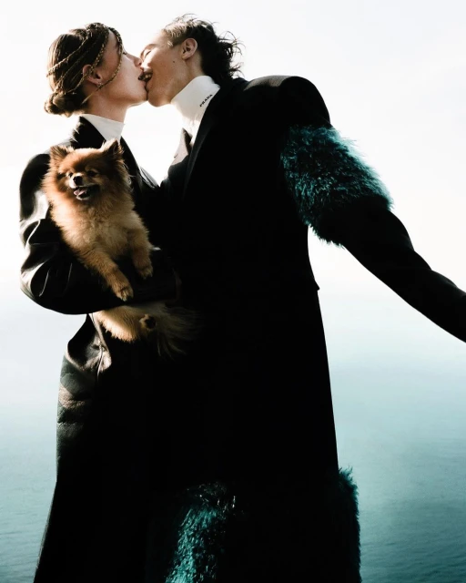 Two people kiss while holding a Pomeranian dog in an outdoor setting with a sea background, showcasing fashion and a touch of nature.