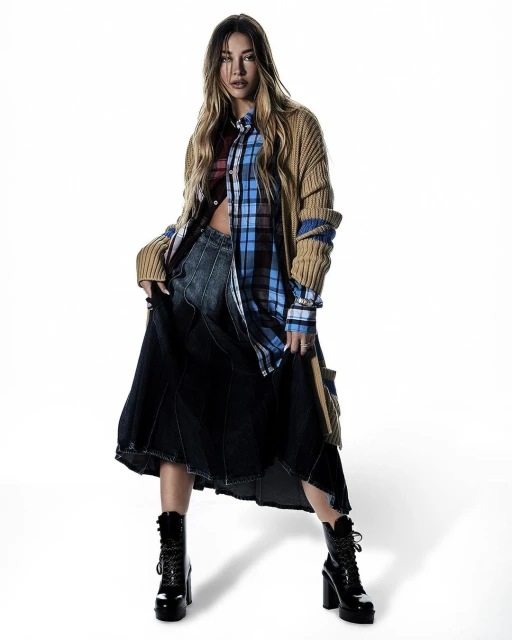 An adult woman is standing in a photo studio wearing a long tan cardigan, plaid shirt, a dark denim skirt, and lace-up heeled boots.
