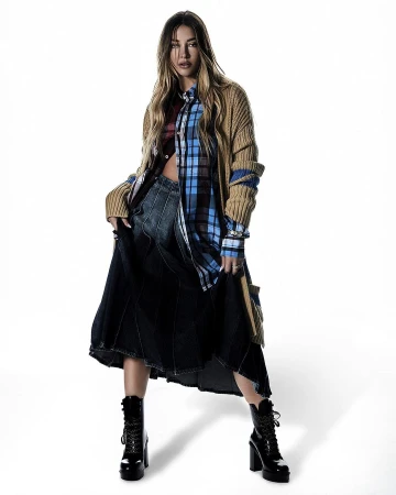 An adult woman is standing in a photo studio wearing a long tan cardigan, plaid shirt, a dark denim skirt, and lace-up heeled boots.