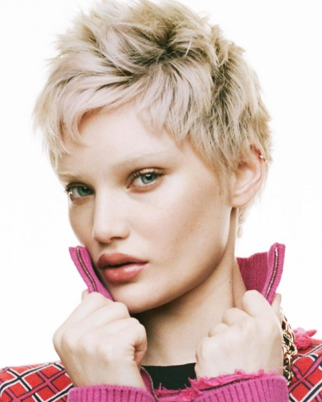 Close-up portrait of a blonde adult female model with blue eyes and pink lips, wearing a red jacket, against a bright white background.