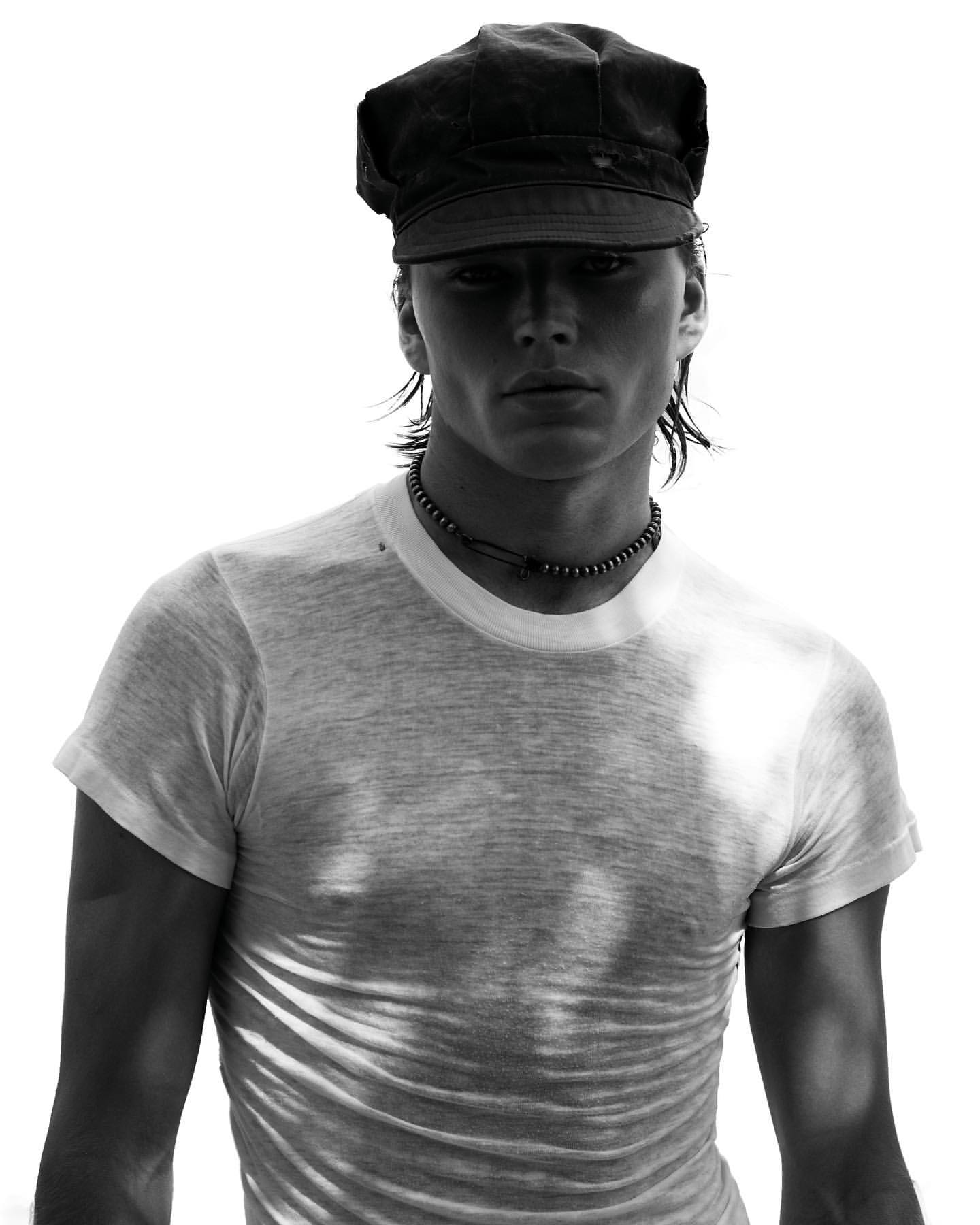 Portrait of a young adult man wearing a white t-shirt and dark cap against a white background, casting shadows on the shirt.