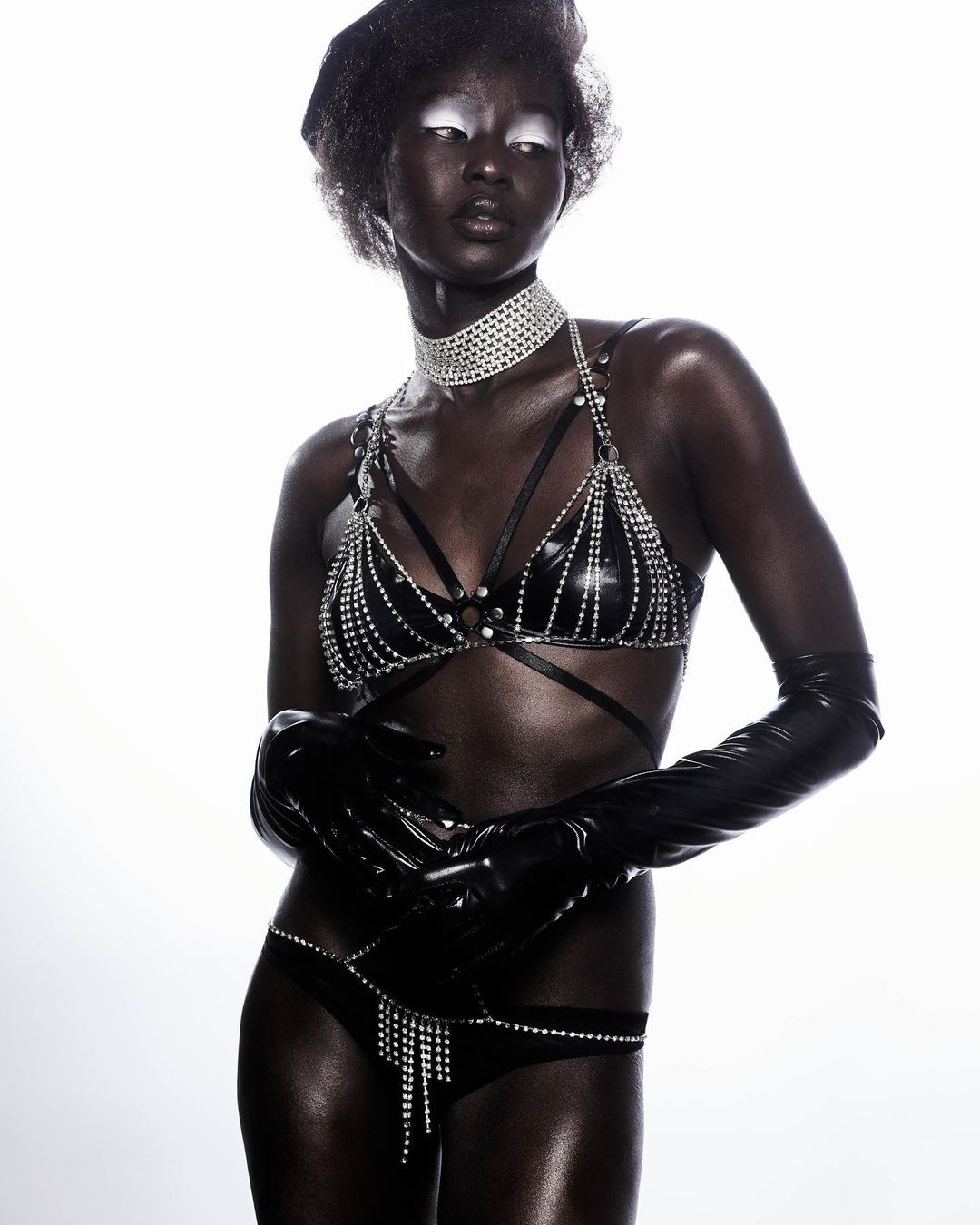 A striking studio portrait of a Black woman, wearing a jeweled black bra and bottom set, accessorized with a choker and long gloves, against white.