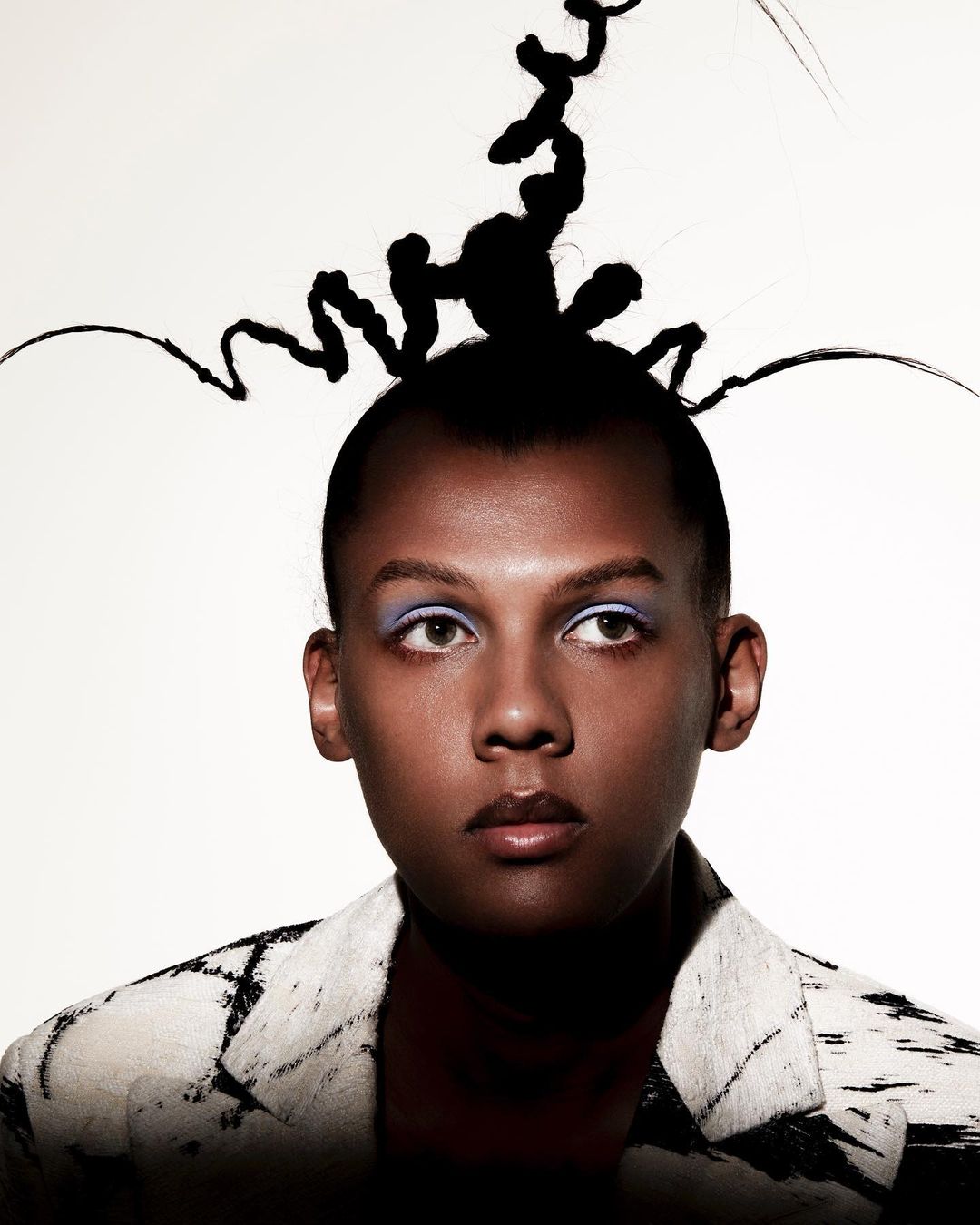 Close-up portrait of a person with dramatic makeup and avant-garde hair, wearing a patterned jacket against a bright background.