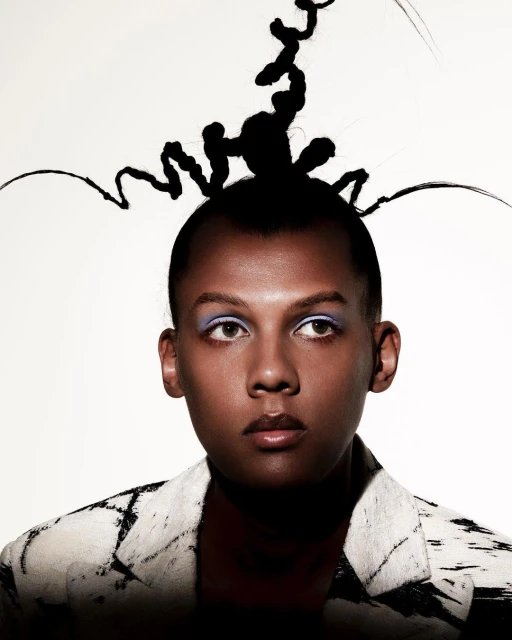 Close-up portrait of a person with dramatic makeup and avant-garde hair, wearing a patterned jacket against a bright background.