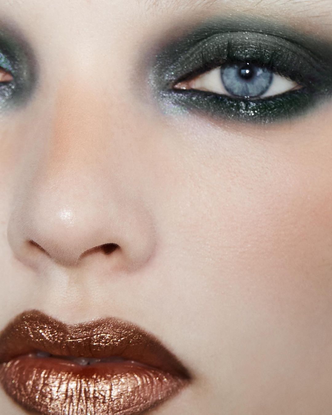 Close-up portrait of a fair-skinned adult woman with blue eyes, dark eyeshadow, and glossy bronze lipstick in an indoor setting.