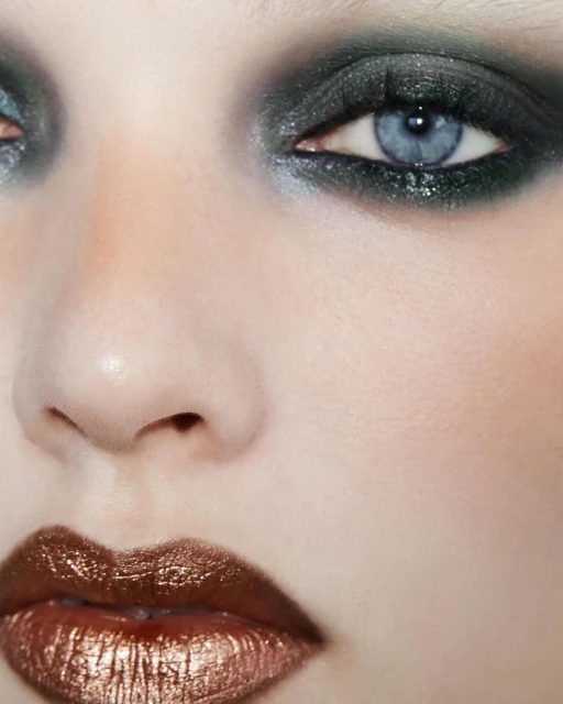 Close-up portrait of a fair-skinned adult woman with blue eyes, dark eyeshadow, and glossy bronze lipstick in an indoor setting.