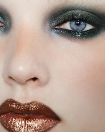 Close-up portrait of a fair-skinned adult woman with blue eyes, dark eyeshadow, and glossy bronze lipstick in an indoor setting.