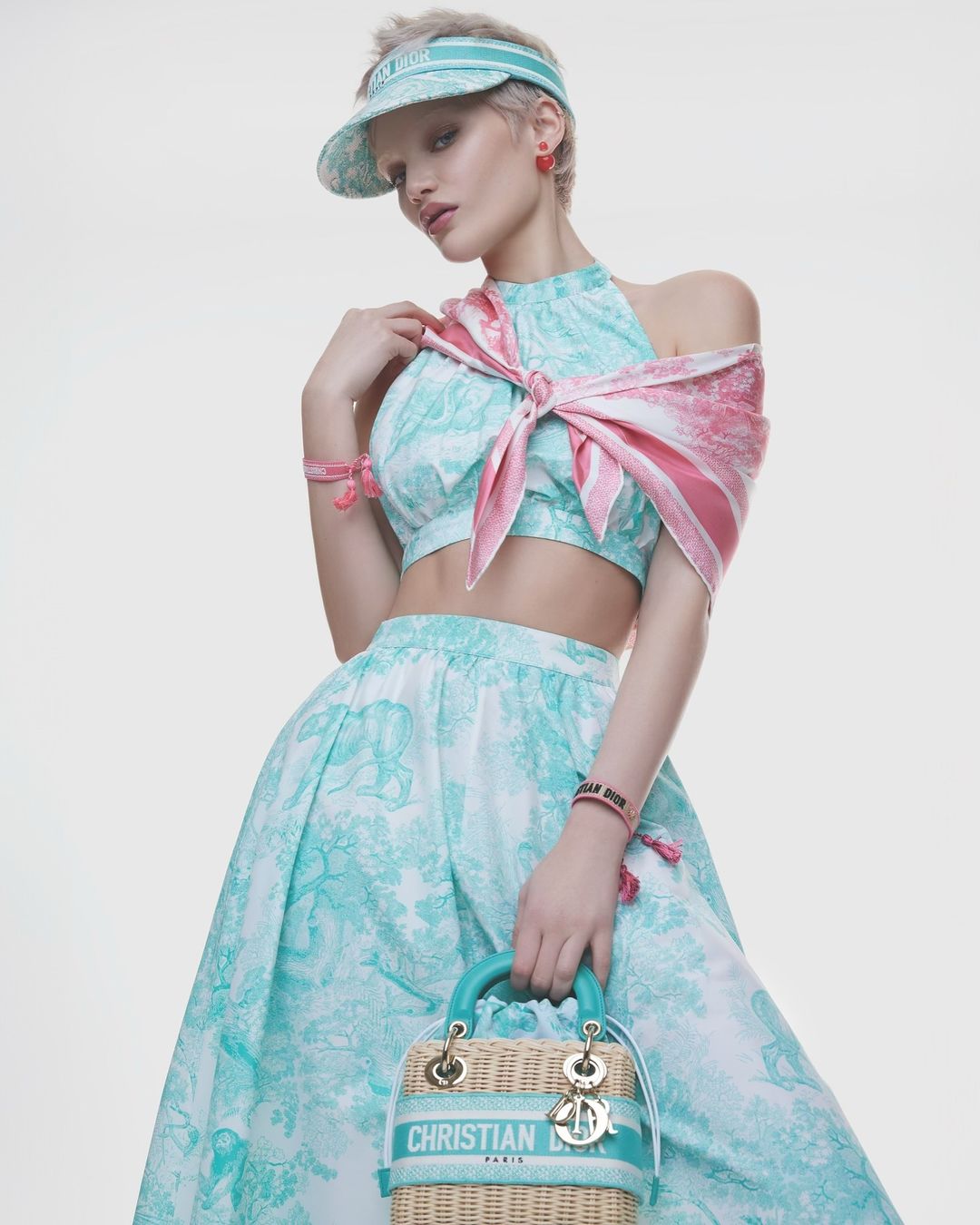 Model in mint and white patterned outfit wearing visor hat holding wicker handbag against white backdrop