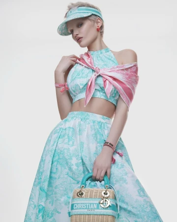 Model in mint and white patterned outfit wearing visor hat holding wicker handbag against white backdrop