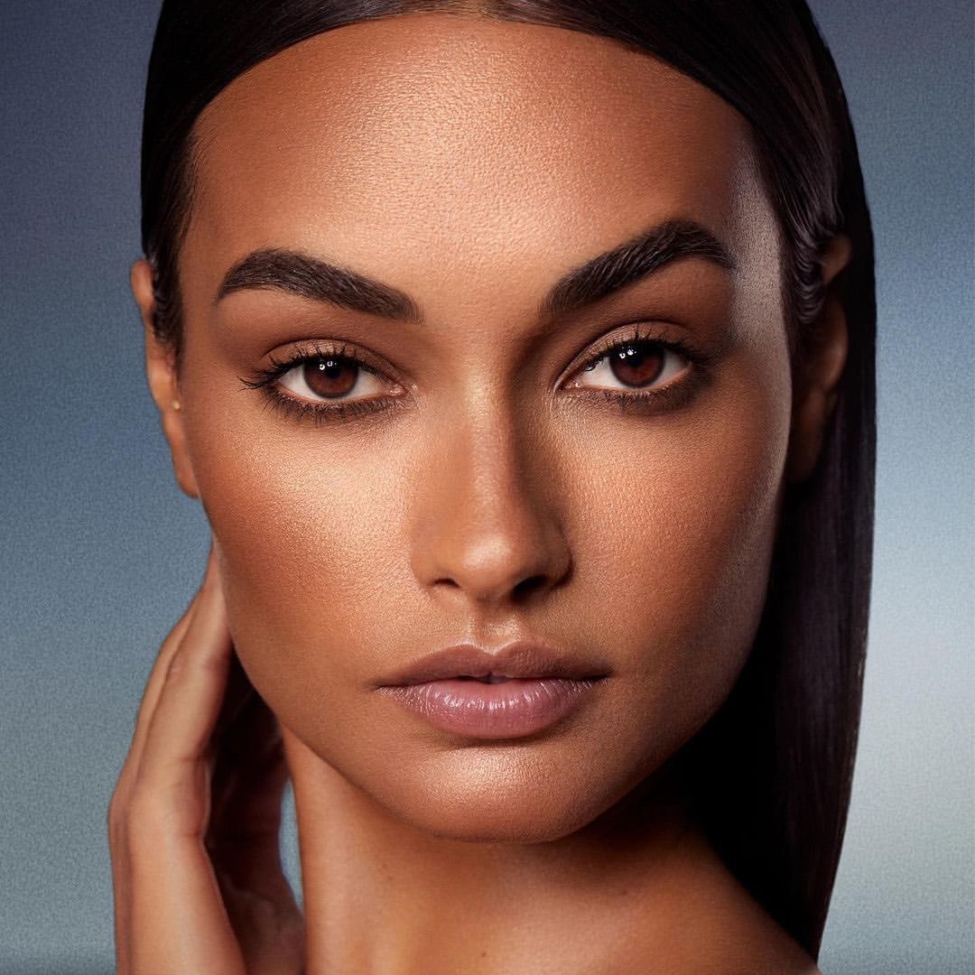 Close-up portrait of a woman with sleek dark hair and glowing skin, her hand gently touching her neck against a muted gradient background.