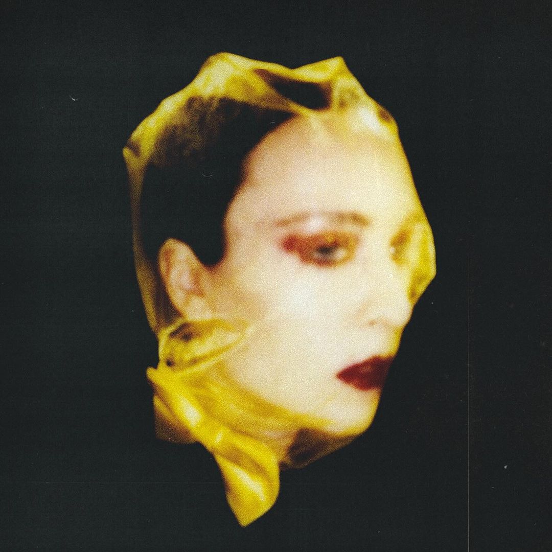 Headshot of woman with dark red lipstick covered in yellow translucent fabric against black backdrop. Studio lighting, high fashion editorial.