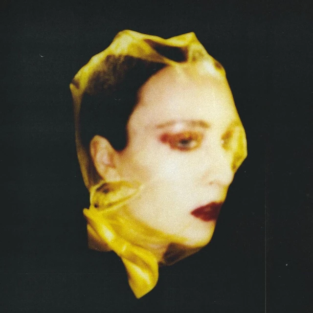 Headshot of woman with dark red lipstick covered in yellow translucent fabric against black backdrop. Studio lighting, high fashion editorial.