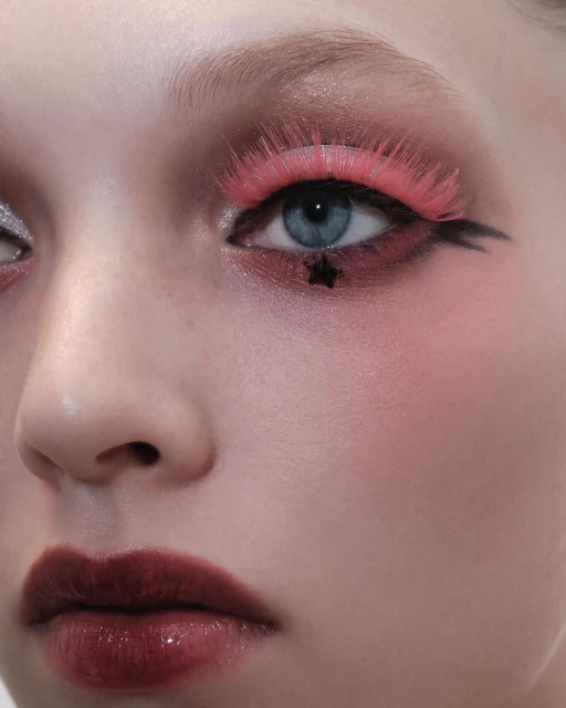 Close-up portrait of a young woman with dramatic pink eye makeup, dark lipstick, and a star detail on her face in a studio setting.