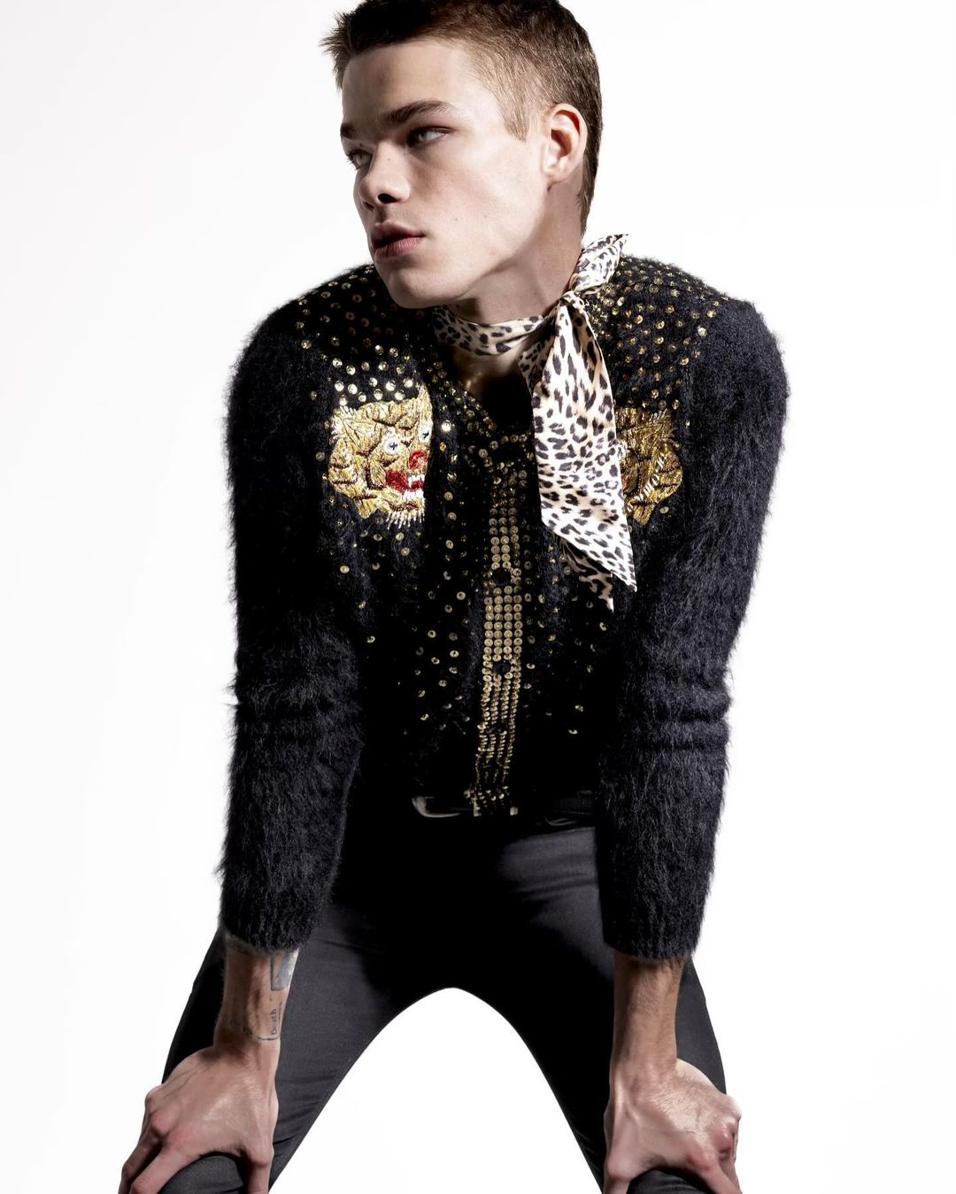 Portrait of a young adult male with dark blond hair wearing an ornate black cardigan with gold embellishments and a patterned scarf.  