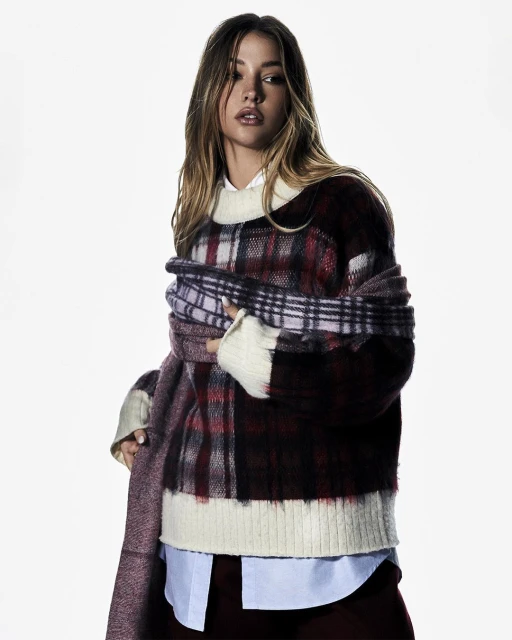 A woman in a plaid sweater and scarf poses in a studio with a white background, looking off to the side.