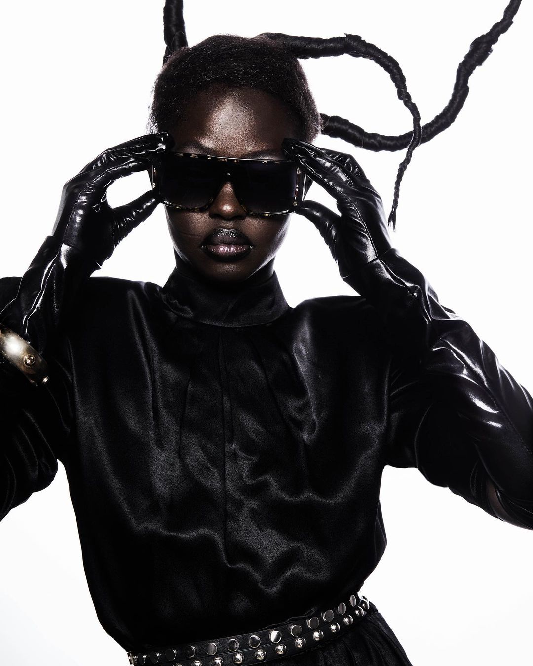 Fashion shot of an adult woman wearing sunglasses and black gloves, striking a pose against a white backdrop with dramatic lighting.