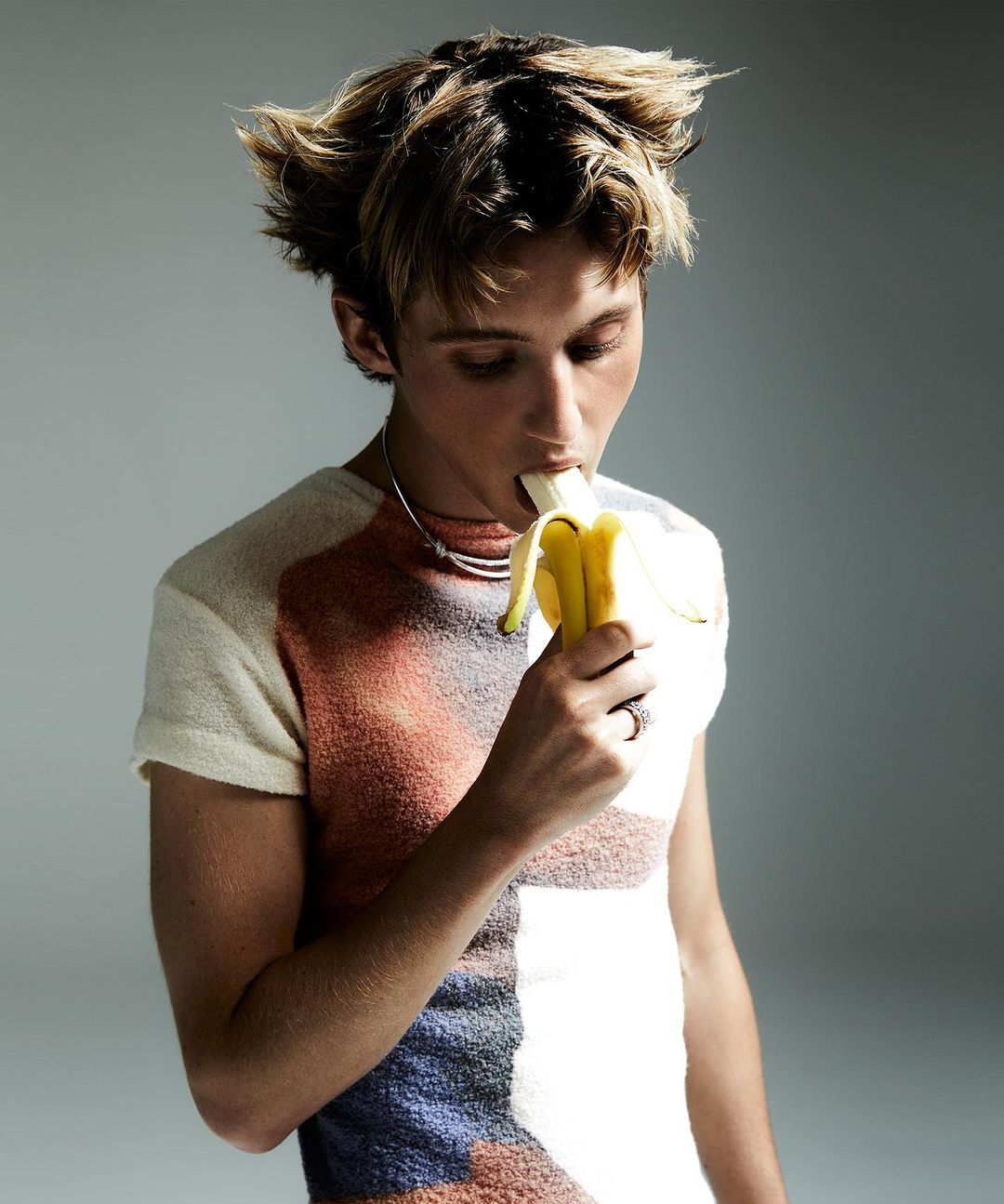 Young adult eats a banana. Blond male holds a yellow fruit, wearing a short-sleeved shirt, and a choker necklace in a studio shot.