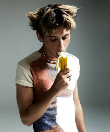 Young adult eats a banana. Blond male holds a yellow fruit, wearing a short-sleeved shirt, and a choker necklace in a studio shot.