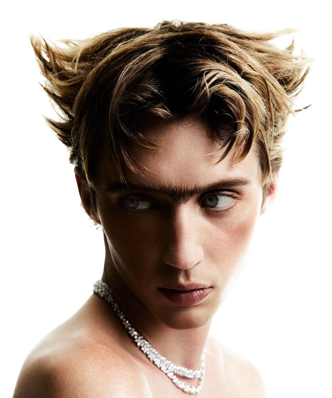 Close-up portrait of a young adult male with unique facial features, tan skin, and light brown hair, wearing a jeweled necklace against a white background.