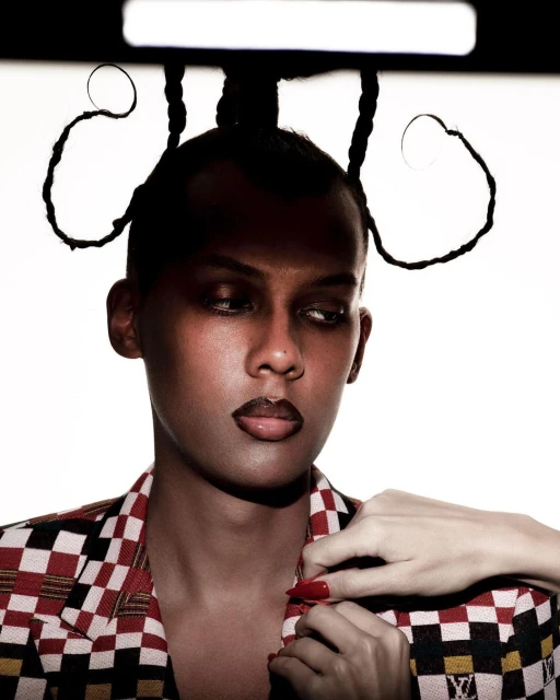 Portrait of an adult male wearing a patterned blazer, dark lipstick, and an elaborate hairstyle with braided extensions, against a white backdrop.