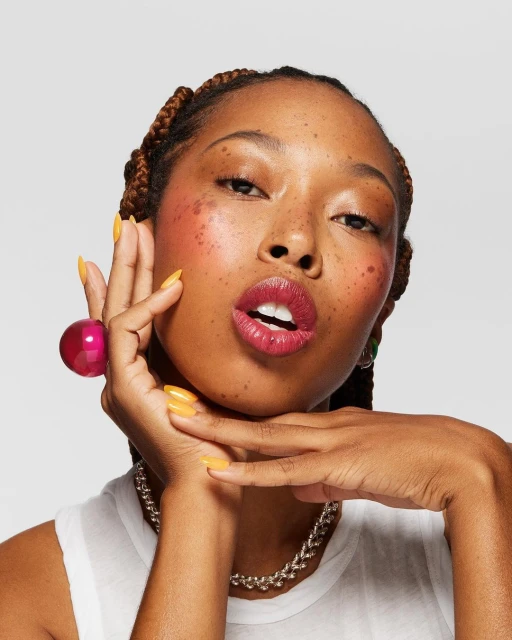 Close-up of a young Black woman in a white top with yellow nails and pink lipstick against a plain white backdrop.
