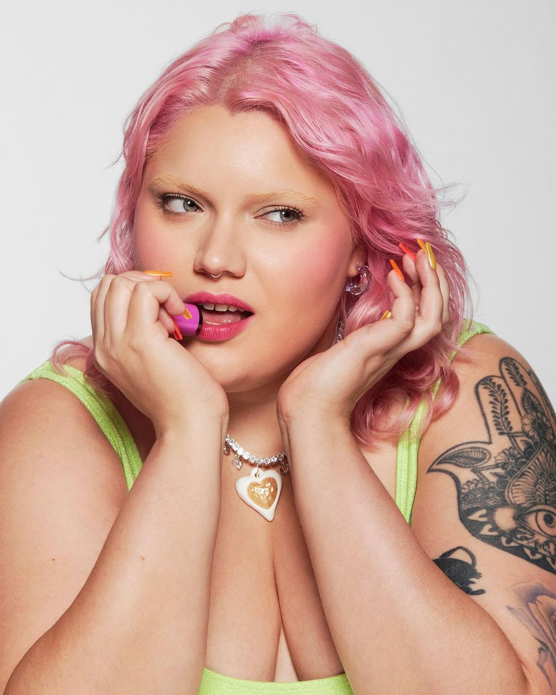 Close-up of a young adult female with pink hair holding a purple makeup sponge next to her face, set against a neutral background.