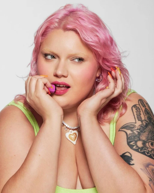 Close-up of a young adult female with pink hair holding a purple makeup sponge next to her face, set against a neutral background.