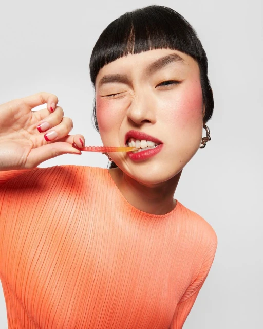 A young Asian woman with dark hair and red lipstick is indoors, posing with a candy stick in her mouth. She is wearing an orange top.