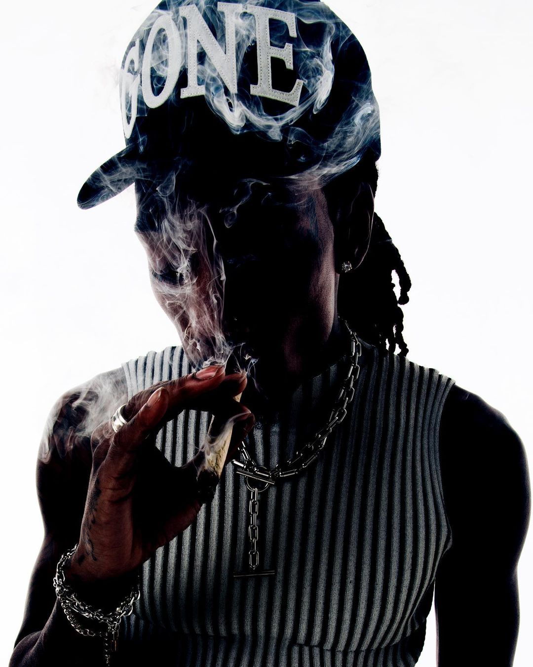 Person smoking a cigarette, wearing a hat with 'GONE' printed on it, silver chain necklaces, with smoke around the face against a white background.