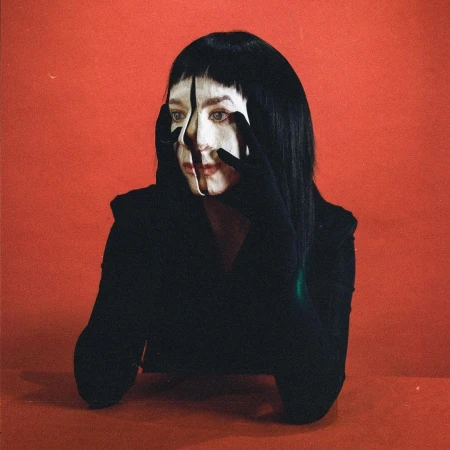 Woman with painted white face and black hair wearing black gloves framing her face against a red background