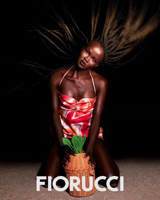 Stunning studio portrait of a bald Black woman in a red and white dress kneeling with a whimsical carrot-shaped woven bag.