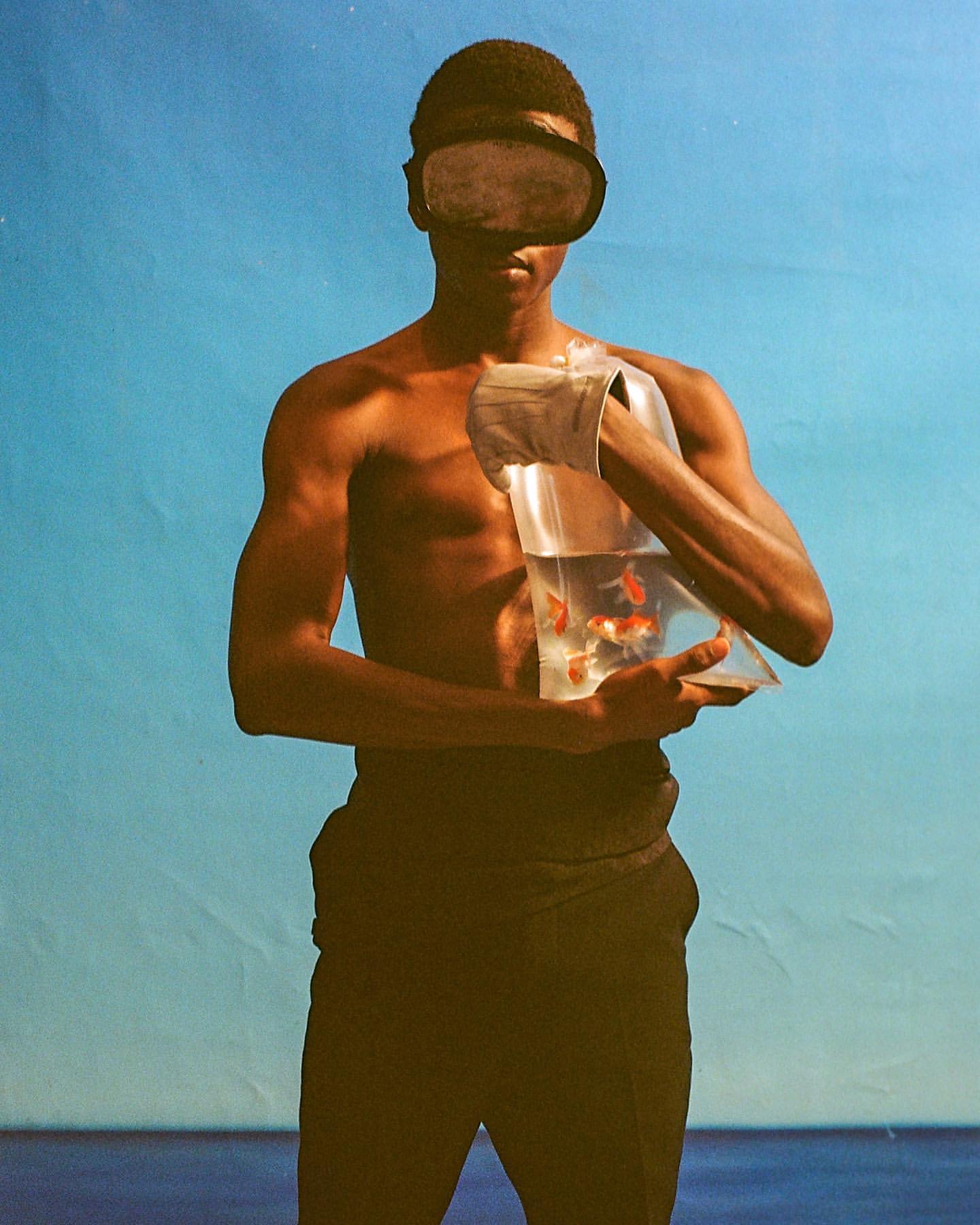 Shirtless black man in sunglasses holds a plastic bag with orange goldfish against a blue sky and horizon in a surreal composition.