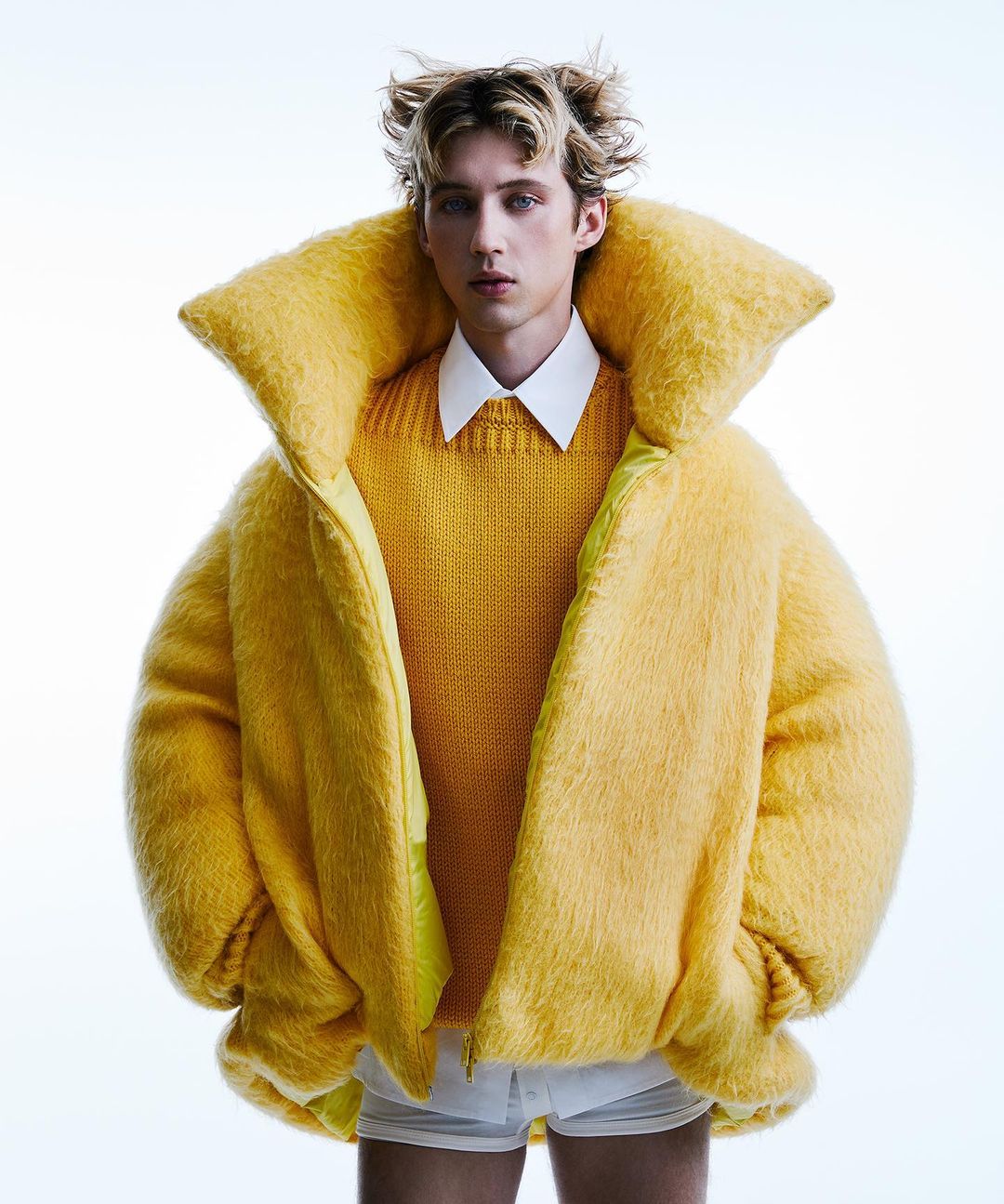 Young male model with blonde hair poses indoors wearing a yellow sweater and oversized fuzzy yellow jacket on a white seamless backdrop.