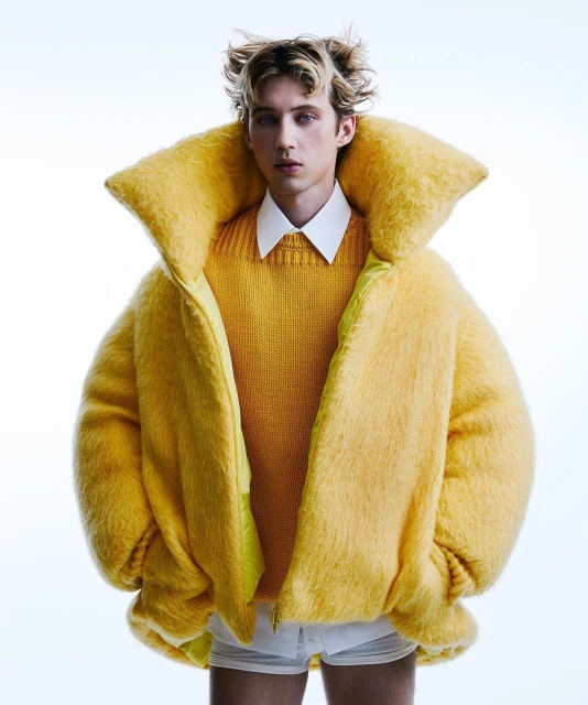 Young male model with blonde hair poses indoors wearing a yellow sweater and oversized fuzzy yellow jacket on a white seamless backdrop.