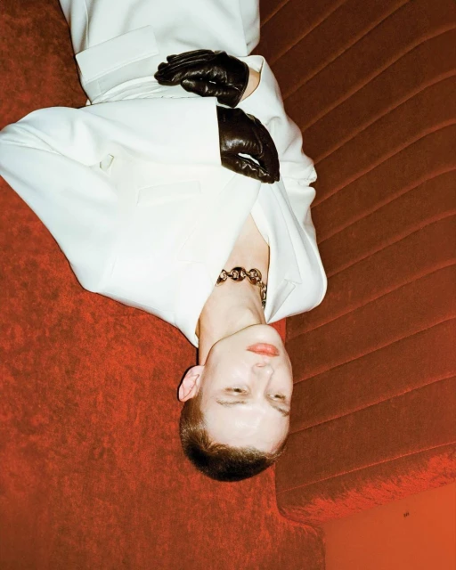 Fashionable person in white suit poses on red velvet sofa with black leather gloves and silver chain necklace in an edgy editorial shot.