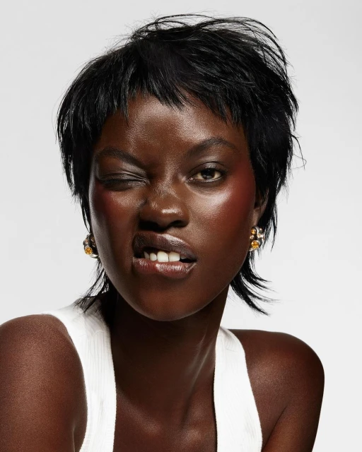 Headshot of an African woman with short black hair, gold earrings, wearing a white tank top, winking, and a light gray background.