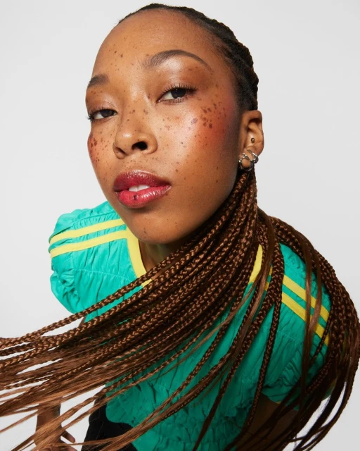 Close-up shot of a woman with brown braided hair and glossy red lipstick posing against a white background wearing a green and yellow top.