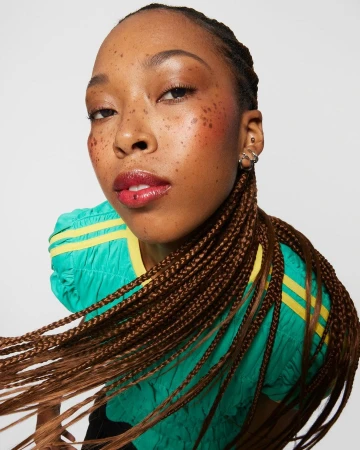 Close-up shot of a woman with brown braided hair and glossy red lipstick posing against a white background wearing a green and yellow top.