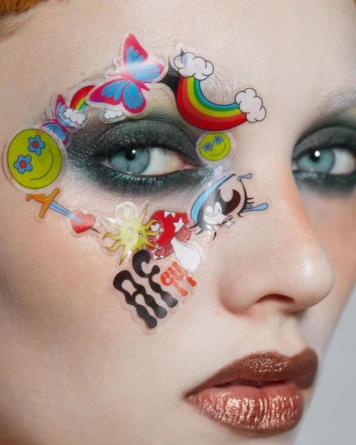 Close-up beauty shot of a fair-skinned woman with colorful cartoon stickers around her eye, smokey eyeshadow, and metallic lips.