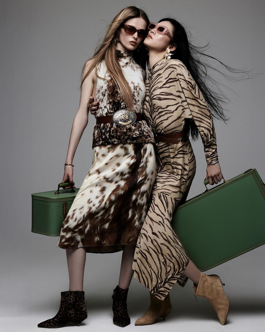 Two women pose in animal-print fashion with vintage green suitcases and neutral-toned boots in a studio setting with muted background.