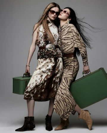 Two women pose in animal-print fashion with vintage green suitcases and neutral-toned boots in a studio setting with muted background.