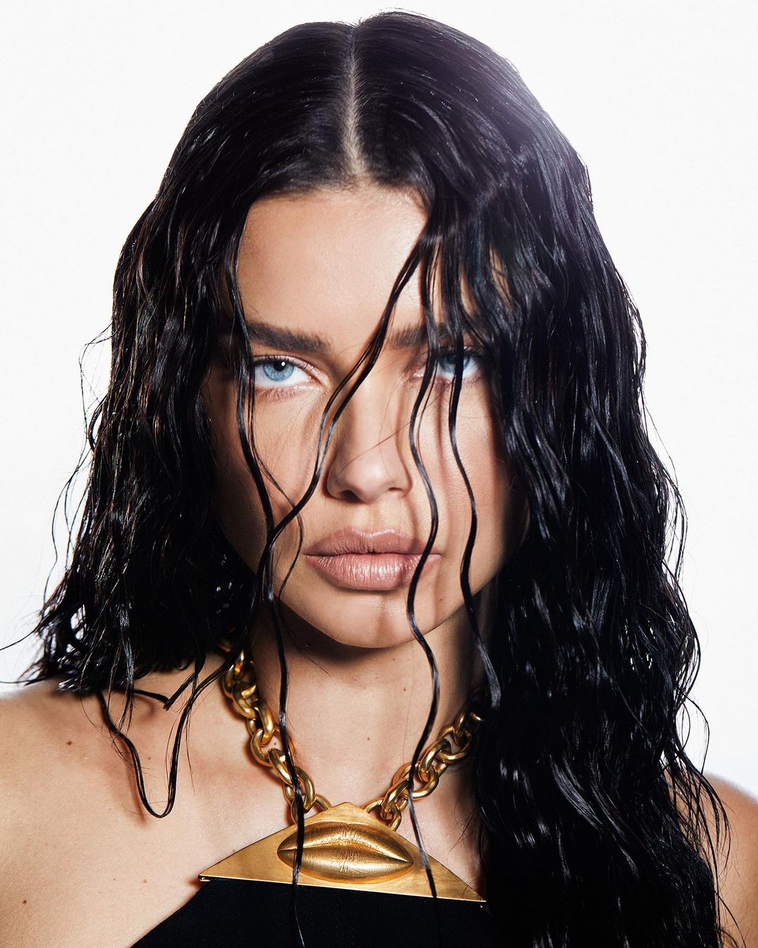 Close-up portrait of an adult female with black wet hair, light eyes, wearing gold necklace against white background, in studio lighting.