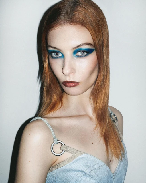 Close-up portrait of a woman with red hair and blue eye makeup, wearing a light blue top against a white background in a beauty shot.