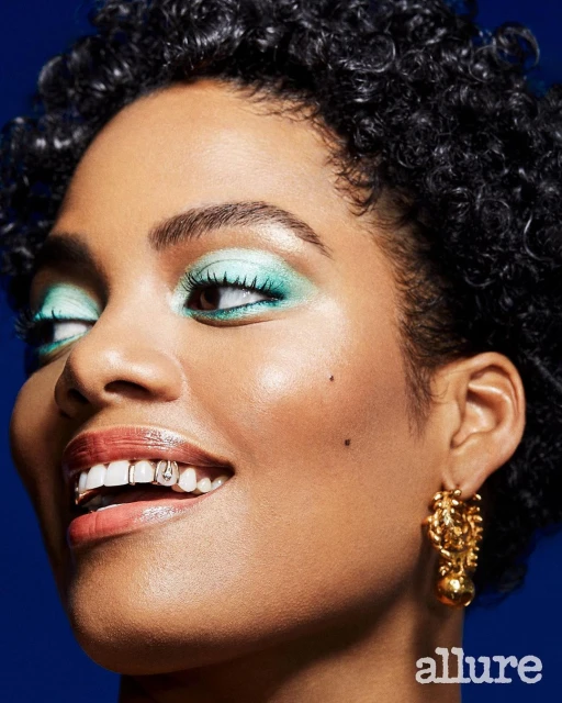 Close-up shot of a smiling adult female model with curly hair, eye shadow, gold earrings, and dental jewelry against a blue backdrop.